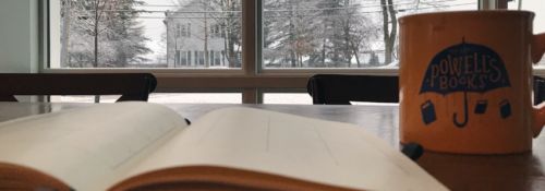 A book and a mug sitting on a table in front of a window overlooking a snowy day